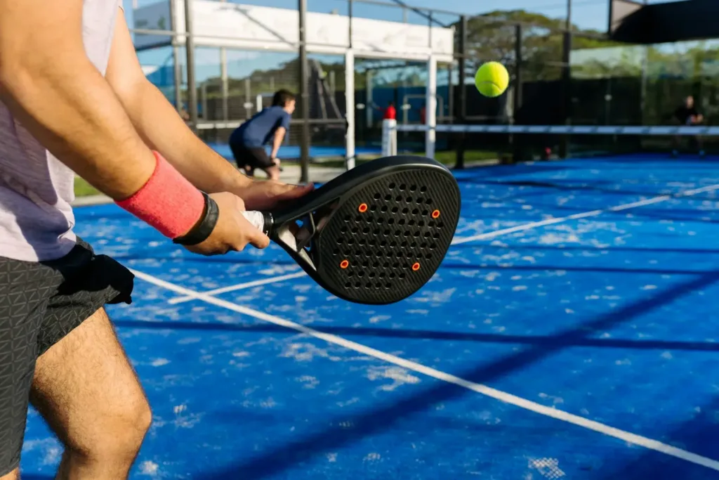 Jogador de padel segurando raquete preta com detalhes laranja em quadra azul, prestes a rebater bola amarela, com outro atleta ao fundo próximo à rede.