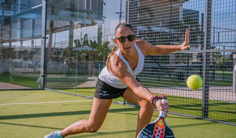 Mulher jogando padel em quadra externa, usando óculos escuros, camiseta branca e shorts pretos com o logo “Rafa Nadal Club”, em posição de ataque para rebater uma bola amarela com raquete preta e vermelha, sob luz natural.