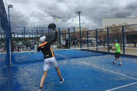 Partida de padel em quadra azul ao ar livre, com quatro jogadores em ação e público assistindo ao fundo sob céu nublado.