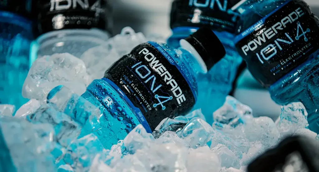 Garrafas de Powerade ION4 sabor Mountain Blast em meio a cubos de gelo, com gotas de condensação destacando o aspecto gelado e refrescante da bebida esportiva azul.