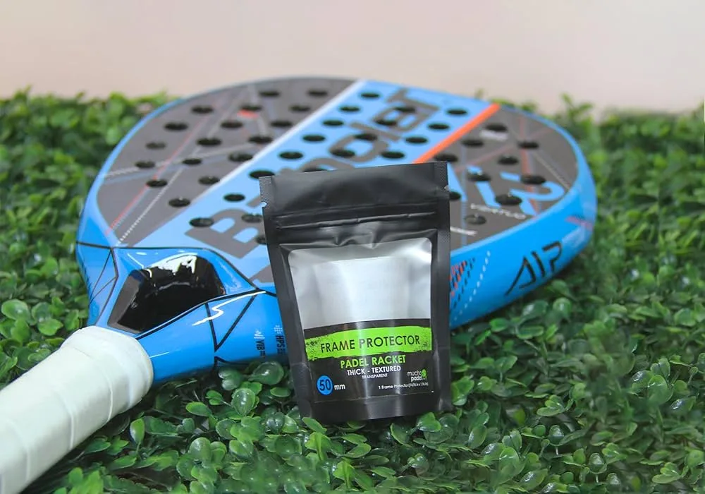 Raquete de padel azul com detalhes em preto e vermelho posicionada sobre folhas verdes artificiais, ao lado de embalagem preta de protetor de quadro (Frame Protector) para raquete, destacando acessórios de proteção esportiva.