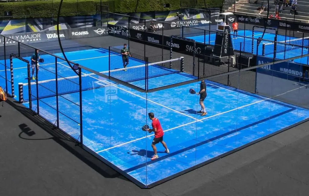 Partida de padel em quadra azul oficial, com quatro jogadores em duplas, cercada por vidro e estrutura metálica, durante torneio profissional.