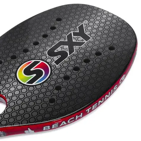 Close-up de uma raquete de beach tennis preta com textura hexagonal, furos simétricos e logotipo colorido em forma de “S”. A borda vermelha exibe a inscrição “BEACH TENNIS” em letras brancas.