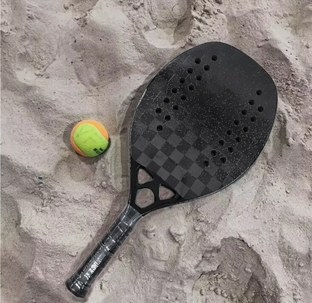 Raquete de beach tennis preta com padrão quadriculado e furos simétricos, posicionada sobre areia clara ao lado de uma bola amarela e laranja, sugerindo ambiente de jogo na praia.