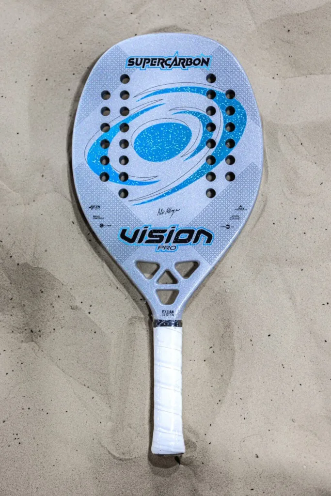 Raquete de beach tennis Vision Pro SuperCarbon sobre areia, com design italiano em tons de prata e azul, mostrando furos simétricos na face e cabo branco envolto em grip.