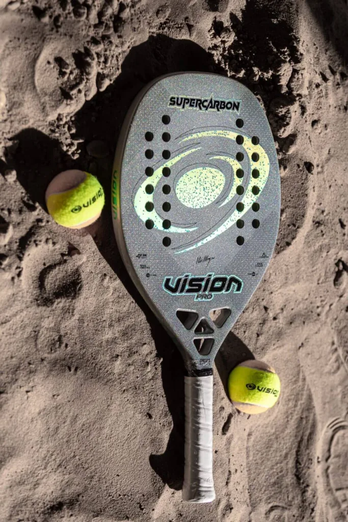 Raquete de beach tennis Vision Pro SuperCarbon sobre areia, acompanhada de duas bolas amarelas da marca Vision, destacando o design moderno com furos simétricos e acabamento metálico.