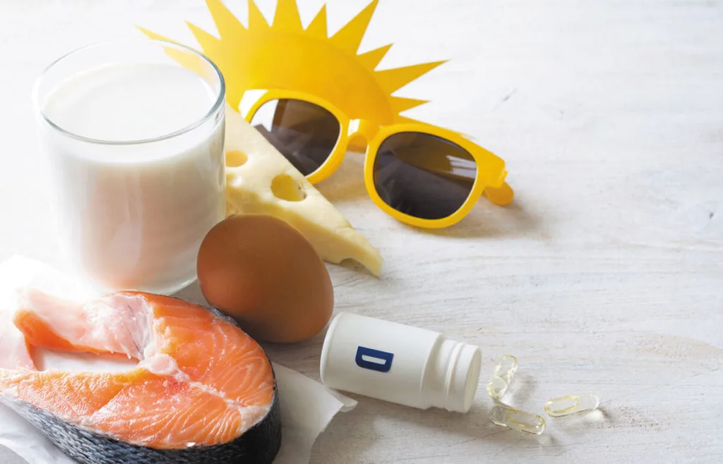 Alimentos ricos em vitamina D — salmão, ovo, queijo e leite — dispostos sobre uma superfície clara, acompanhados de cápsulas de suplemento e óculos de sol amarelos com recorte de sol ao fundo, simbolizando a importância da exposição solar.