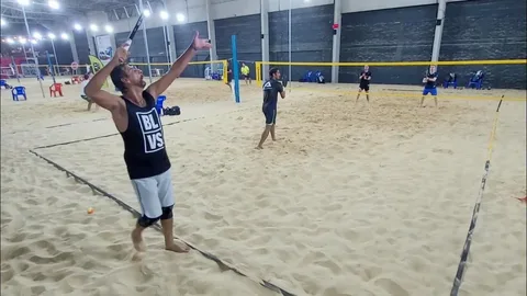 Jogadores disputando uma partida de beach tennis indoor, em quadra de areia cercada por paredes e iluminação artificial, com um atleta em primeiro plano preparando o saque e outros três posicionados do outro lado da rede.