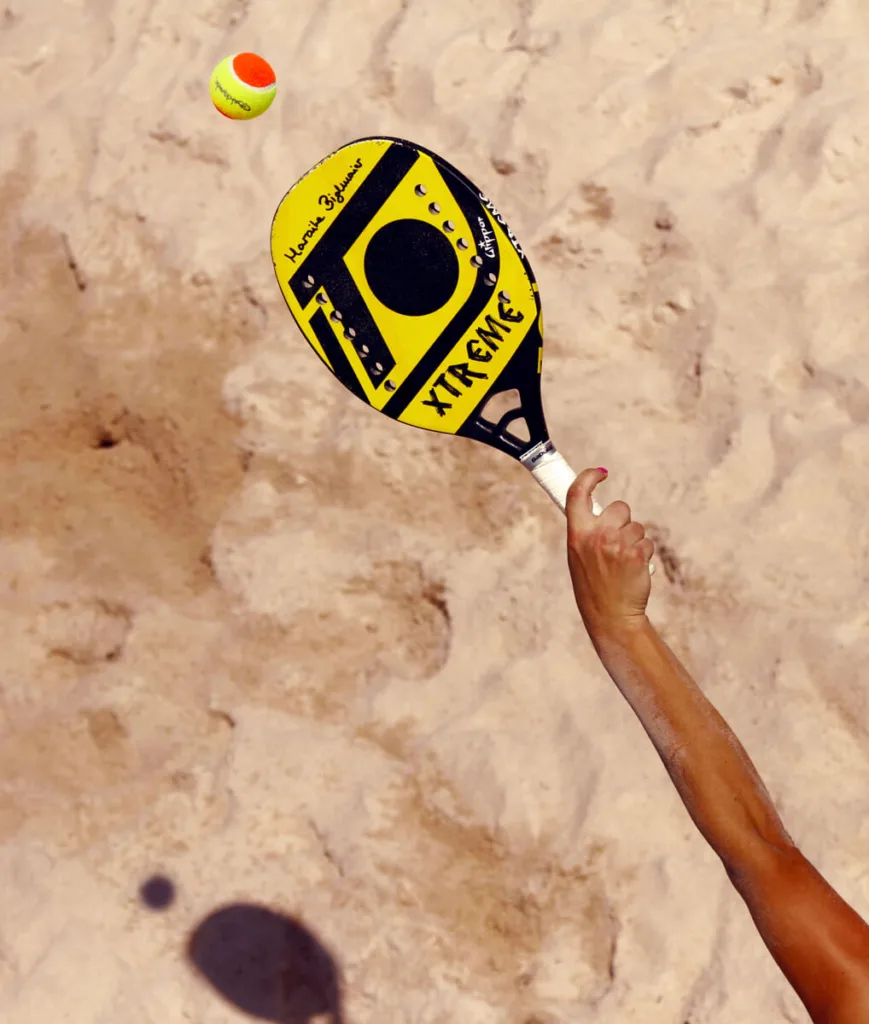 Raquete de padel Babolat e quatro bolas amarelas sobre quadra verde de grama sintética, posicionadas próximas à rede e linha branca lateral, sugerindo início ou pausa de jogo.