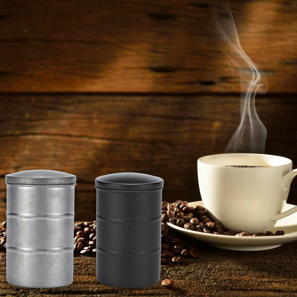 Xícaras de café branco com bebida quente e fumegante ao lado de dois potes metálicos — um prateado e outro preto — sobre mesa de madeira rústica, com grãos de café espalhados ao redor.