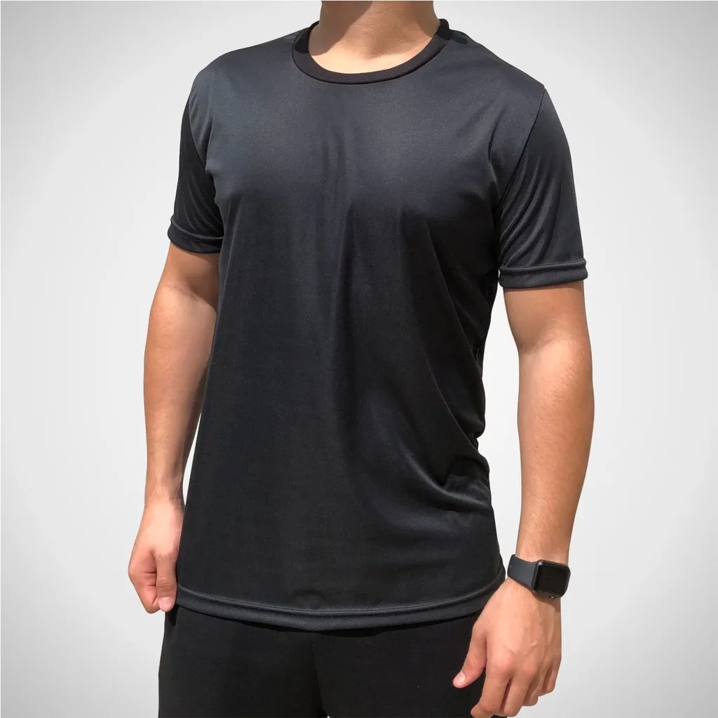 Homem vestindo camiseta preta de manga curta e calça preta, em fundo cinza neutro. A camiseta tem corte simples e tecido com leve brilho, sugerindo material esportivo dry-fit.