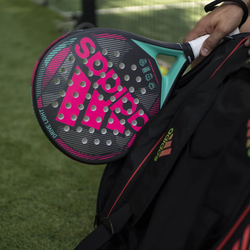 Pessoa segurando raquete de padel Adidas Drive Light preta com detalhes em rosa e azul, acompanhada de bolsa esportiva Adidas preta com detalhes vermelhos e verdes, em quadra com grama ao fundo.