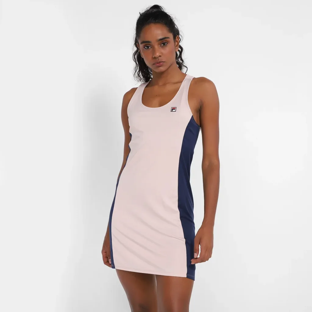 Mulher vestindo vestido esportivo sem mangas da marca Fila, em tom rosa claro com painéis laterais azul-marinho, posando contra fundo branco. O vestido tem corte justo e logo Fila pequeno no peito esquerdo.