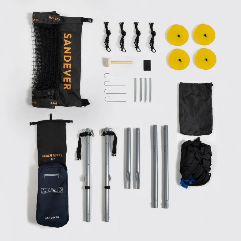 Kit completo de beach tennis Sandever disposto sobre superfície clara, incluindo rede, postes metálicos, cordas, estacas, discos amarelos, martelo e bolsas de transporte pretas com logotipo da marca.