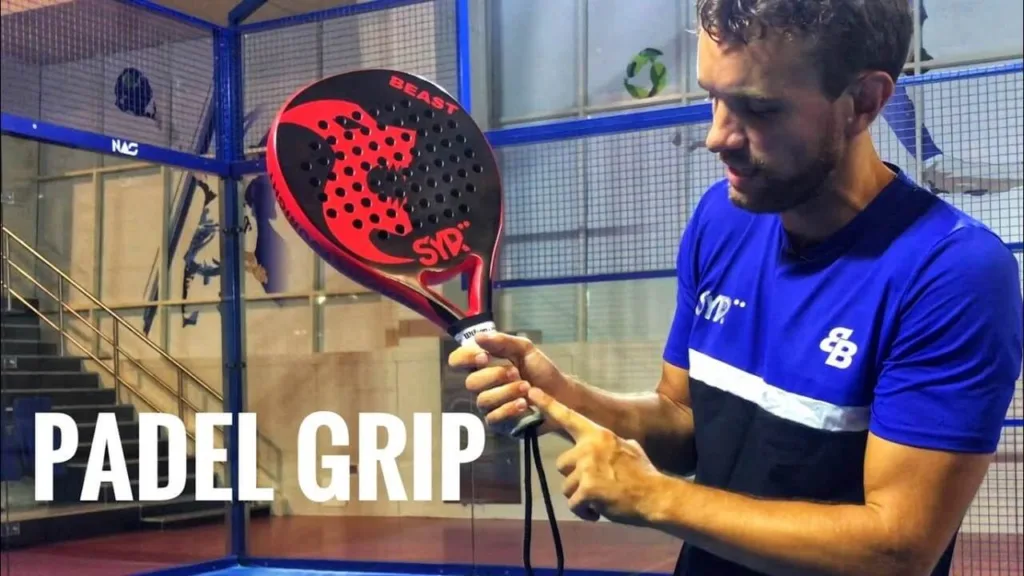 Homem em quadra de padel segurando uma raquete preta e vermelha da marca SVP com o nome “Beast”, demonstrando a forma correta de segurar o grip, vestindo camiseta azul e preta com logotipos esportivos, em ambiente indoor com estrutura metálica e vidro.