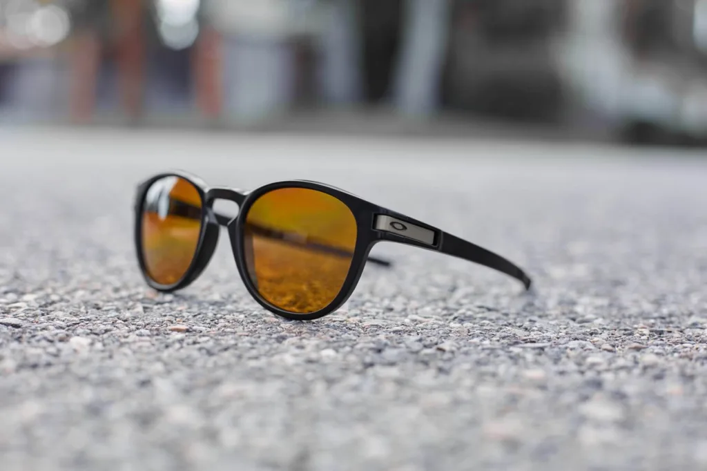 Óculos de sol Oakley com armação preta e lentes espelhadas alaranjadas, posicionados sobre uma superfície de asfalto. O fundo está desfocado, destacando o design moderno e esportivo do acessório.