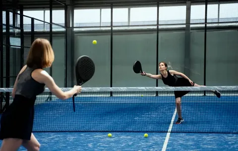 Duas mulheres jogando padel em quadra indoor azul, separadas pela rede, ambas segurando raquetes pretas e focadas na bola amarela em movimento; o ambiente é moderno, com paredes de vidro e estrutura metálica.