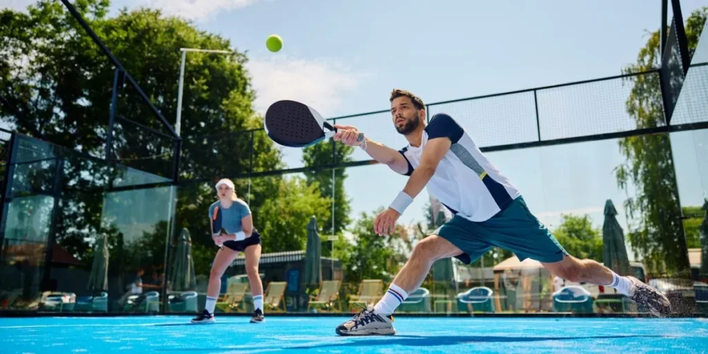 Dois jogadores em quadra de padel azul ao ar livre, um deles avançando para rebater a bola com raquete preta, cercados por paredes de vidro e cenário com árvores ao fundo.