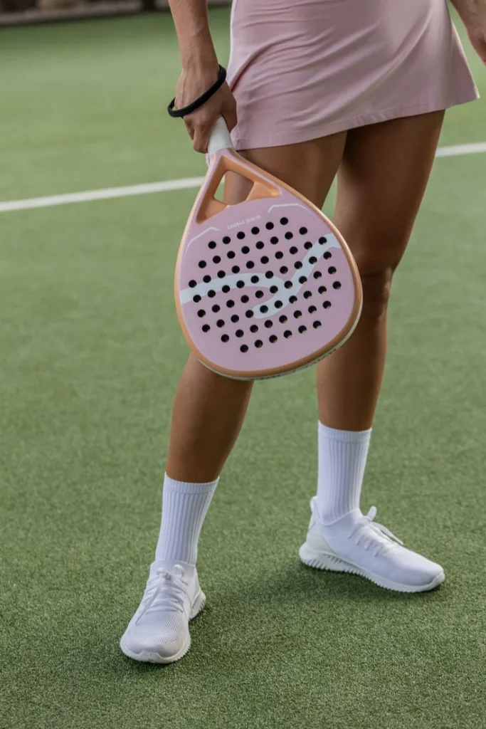 Jogadora em quadra verde segurando raquete de padel rosa em formato lágrima, vestindo saia rosa clara, meias brancas e tênis esportivos brancos.