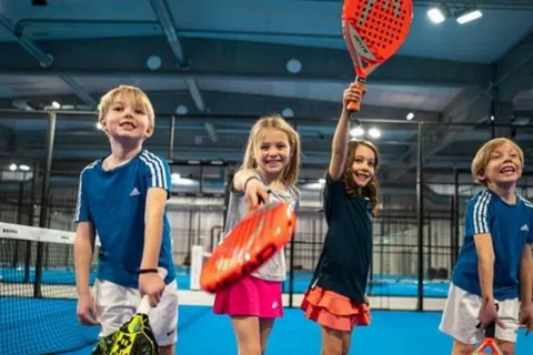 Três crianças em quadra azul de padel segurando raquetes coloridas, posicionadas próximas à rede em ambiente esportivo indoor.