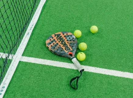 Raquete de padel Babolat e quatro bolas amarelas sobre quadra verde de grama sintética, posicionadas próximas à rede e linha branca lateral, sugerindo início ou pausa de jogo.