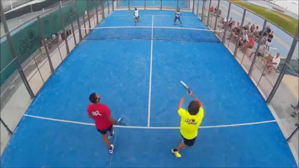 Vista aérea de partida de padel em quadra azul, quatro jogadores em duplas, quadra cercada por vidro e metal, com espectadores assistindo ao jogo.
