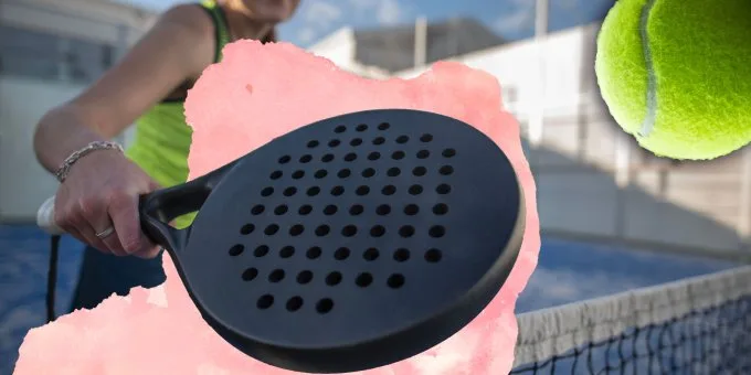 Close-up de raquete de padel preta com furos circulares, segurada próximo à rede em quadra azul, com bola amarela em destaque.