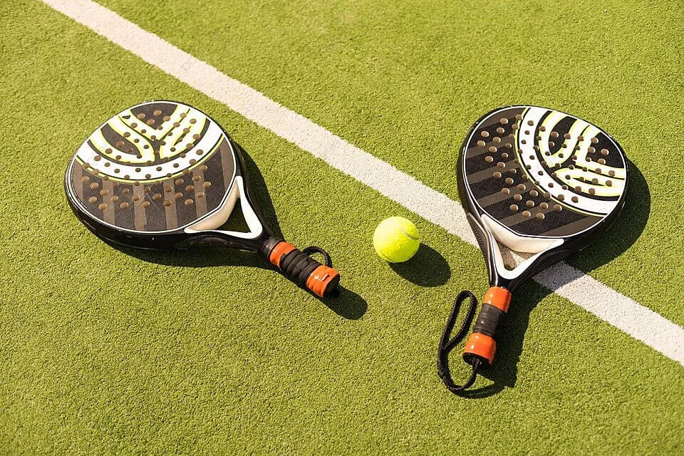 Duas raquetes de padel com design preto e branco e fitas laranja no cabo, acompanhadas de uma bola amarela sobre quadra de grama sintética verde.