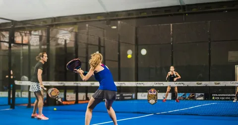 Quatro jogadores em quadra azul de padel, cercada por paredes de vidro, disputando partida em duplas com espectadores assistindo ao fundo.