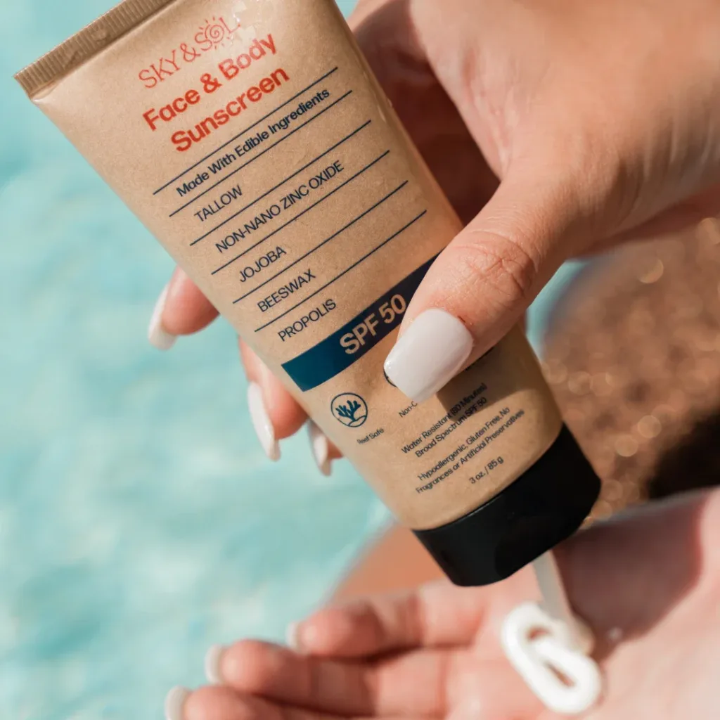 Mão aplicando protetor solar SKY & SOL Face & Body SPF 50, embalagem bege com detalhes em azul e laranja, fundo com piscina.