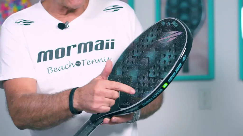 Homem vestindo camiseta branca da marca Mormaii Beach Tennis segura e aponta para uma raquete de beach tennis preta com textura e furos na face, destacando detalhes do design e acabamento.