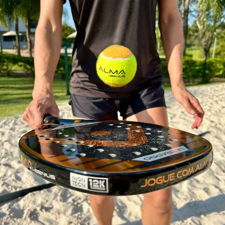 Pessoa segurando uma raquete de beach tennis ALMA Genius com acabamento em carbono 12K, enquanto uma bola amarela com logotipo ALMA Genius flutua em frente à camiseta preta da marca, sobre uma quadra de areia ao ar livre.