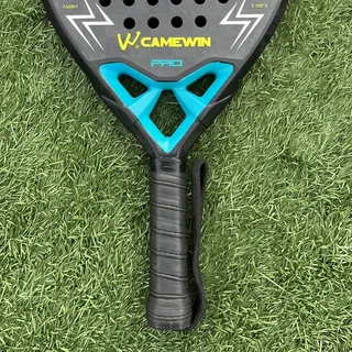 Raquete de padel preta com detalhes azul e turquesa sobre grama sintética, empunhadura com alça de punho e marca Camewin destacada em amarelo.