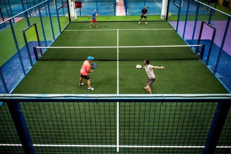 Quatro jogadores em partida de duplas de padel em quadra indoor azul, com rede central e cercado metálico, cada atleta segurando raquete durante o rally.