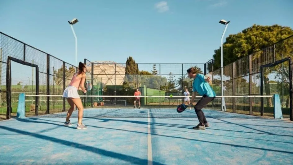Quatro jogadores disputando uma partida de padel em quadra azul ao ar livre, cercada por telas de proteção, com dois atletas em primeiro plano prontos para rebater a bola amarela que cruza a rede.