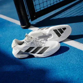 Tênis brancos Adidas com três listras pretas, colocados em uma quadra azul ao lado da rede, mostrando um tênis de perfil e o outro a sola técnica.