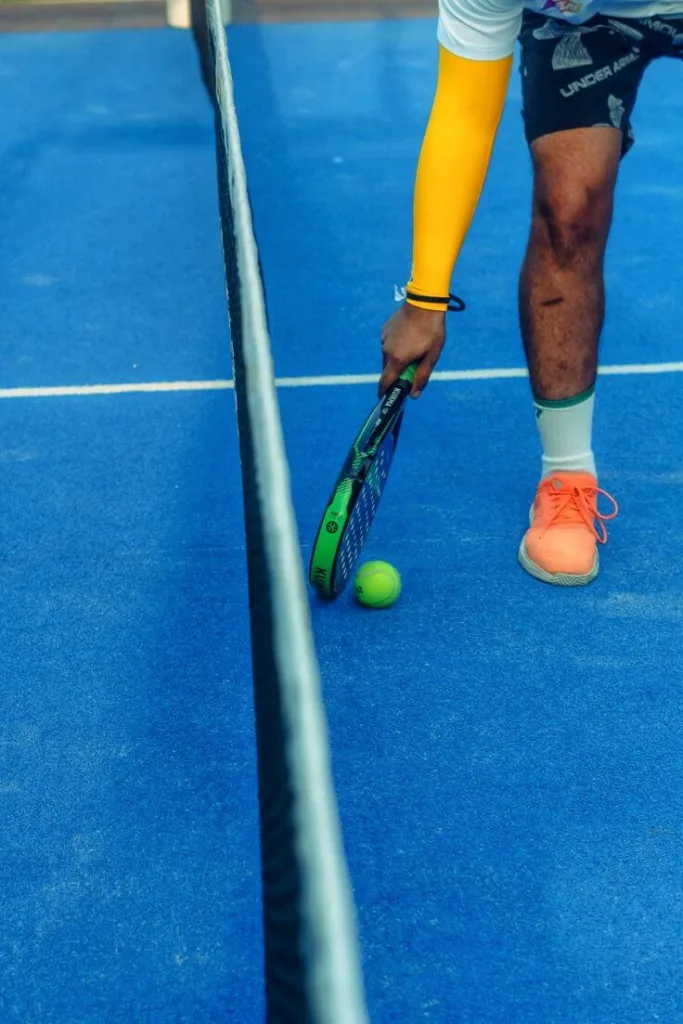 Jogador em quadra azul de padel segurando raquete verde e preta, usando tênis esportivo laranja, prestes a interagir com bola verde próxima à rede.