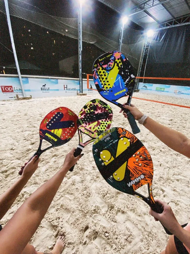 Quatro pessoas segurando raquetes de beach tennis coloridas da marca VAMO sobre uma quadra de areia iluminada à noite, com rede e banners de patrocinadores ao fundo.