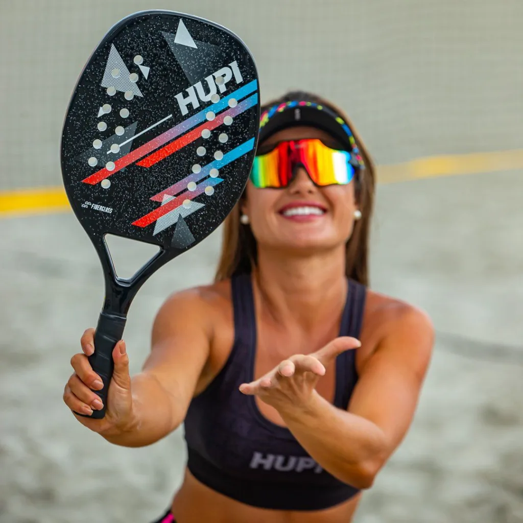 Mulher jogando beach tennis em uma quadra de areia, segurando uma raquete HUPI preta com detalhes geométricos em vermelho, azul e branco. Ela usa viseira preta, top esportivo da marca HUPI e óculos espelhados multicoloridos, sorrindo enquanto lança a bola.