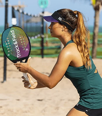 Mulher jogando beach tennis em uma quadra de areia, segurando uma raquete colorida da marca Racket & Led. Ela usa regata verde escura, shorts pretos e viseira preta, com o mar e redes de fundo.