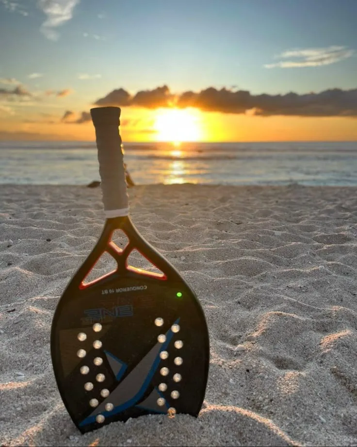 Raquete de beach tennis preta com detalhes em azul e cinza fincada na areia da praia durante o pôr do sol, com o mar e o céu dourado ao fundo.