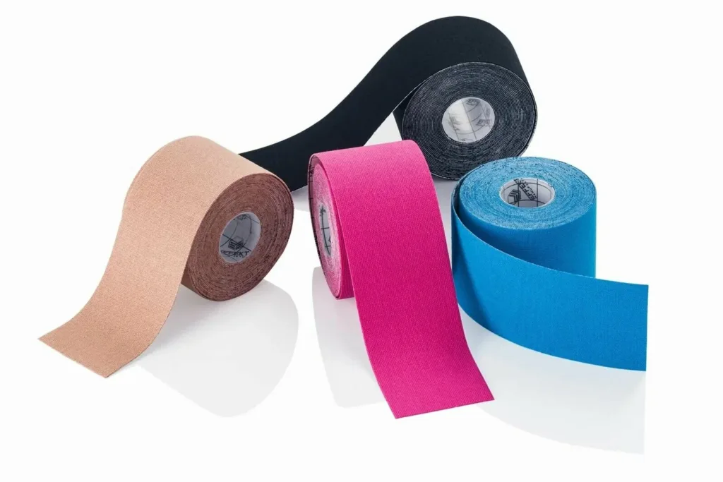 Quatro rolos de fita kinesio terapêutica dispostos sobre uma superfície branca, nas cores bege, preta, rosa e azul, parcialmente desenrolados para mostrar a textura elástica do material.