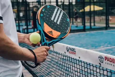 Pessoa segurando uma raquete de padel Adidas e uma bola amarela próxima à rede de uma quadra azul, com o logotipo “Padel Academy” visível na faixa da rede e jogadores ao fundo.