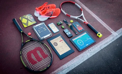 Conjunto de equipamentos esportivos e tecnológicos organizados sobre uma quadra de tênis, incluindo duas raquetes Wilson, tênis Adidas laranja, dispositivos eletrônicos como tablets, smartphones e sensores de desempenho, além de uma bola de tênis.