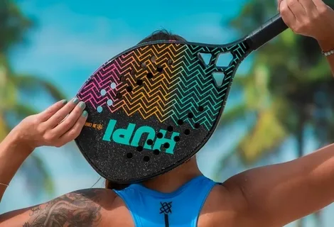 Mulher segurando uma raquete de beach tennis HUPI colorida à frente do rosto, com design em zigue-zague nas cores rosa, laranja e azul sobre fundo preto, em cenário de praia com palmeiras e céu azul ao fundo.