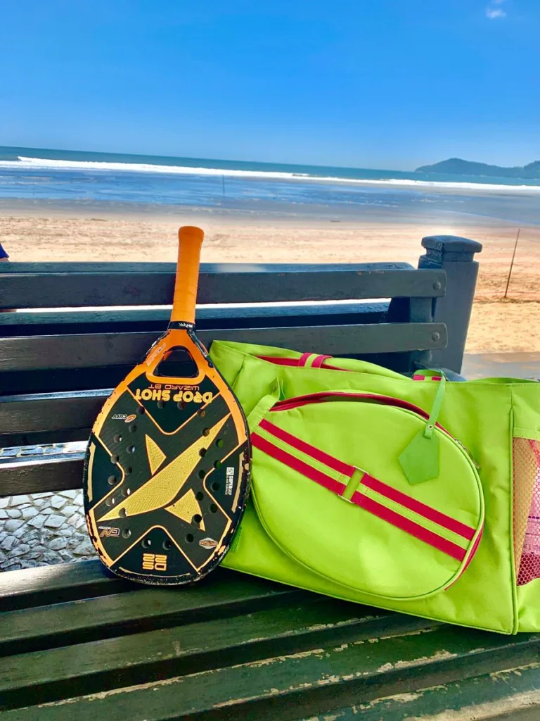 Raquete de beach tennis Drop Shot preta e amarela apoiada em um banco de madeira, ao lado de uma bolsa esportiva verde com detalhes vermelhos. Ao fundo, uma praia com areia clara, mar calmo e céu azul.